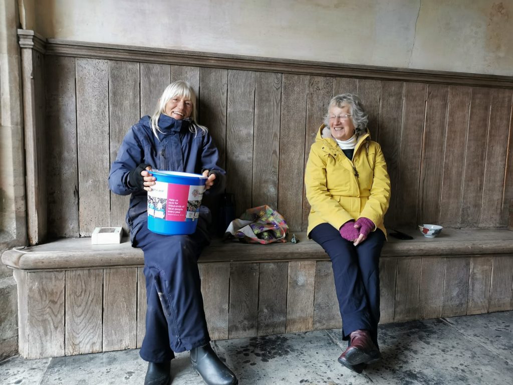 Two women sat on a wooden bench holding a collecting bucket for Hospiscare