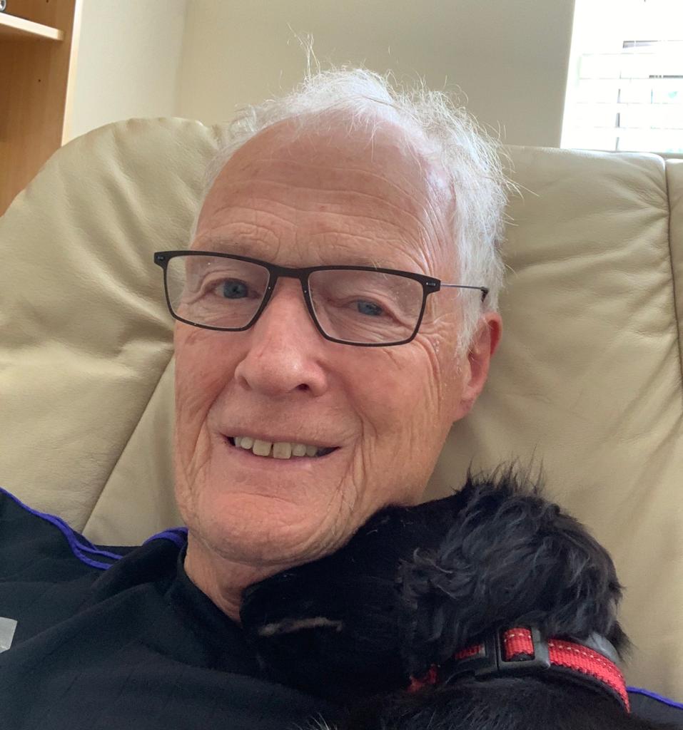 An older man in a chair with a black dog cuddled under his neck
