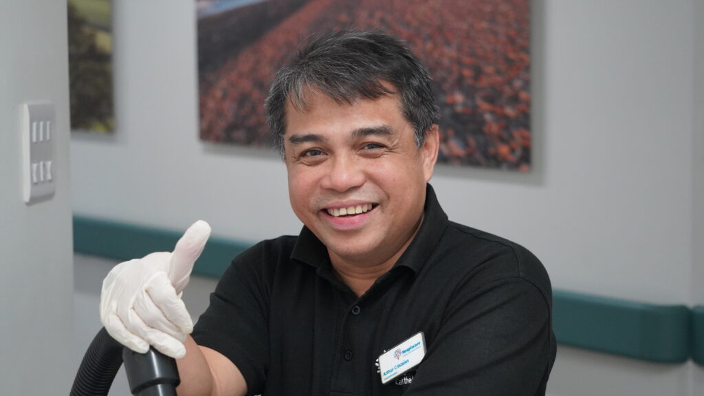 A man smiling at the camera wearing a black polo, a name badge and white latex gloves
