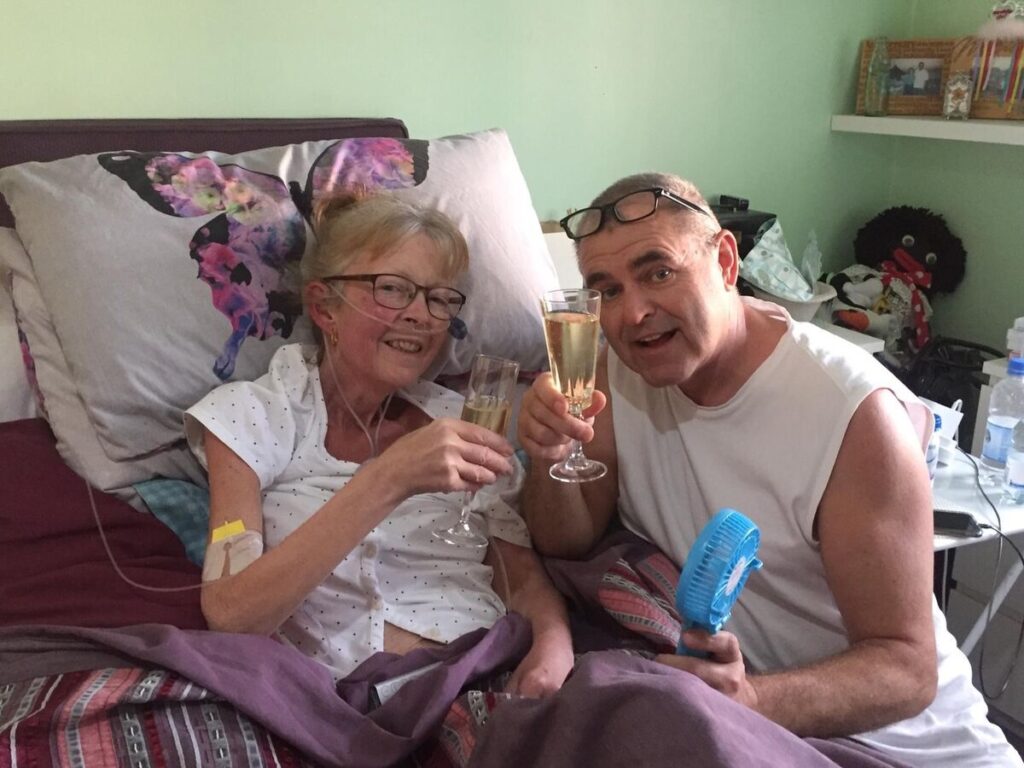Female hospice patient sat in bed holding a glass of champagne to toast her daughter's birthday. Next to her is her husband, also holding champagne. Both are smiling at the camera.