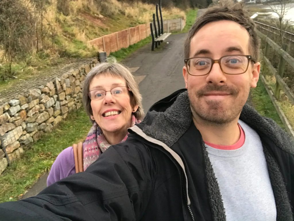 A man with glasses taking an outdoor selfie with a woman smiling behind him